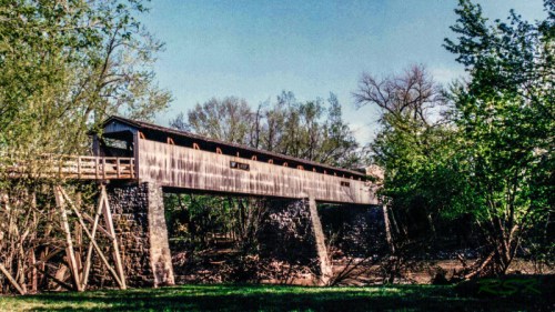 A wooden covered bridge spans a stream, surrounded by lush greenery and trees under a clear blue sky.