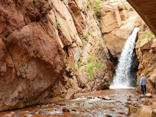 A person stands near a waterfall cascading down rocky cliffs, surrounded by lush greenery and a flowing stream.
