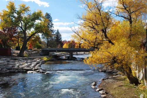 A serene river flows under a bridge, surrounded by vibrant autumn trees and a clear blue sky.