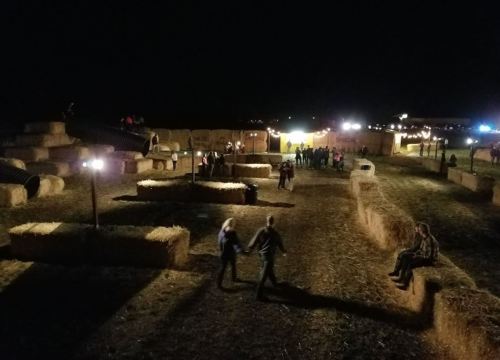 A nighttime scene at a farm with hay bales, people walking, and lights in the background creating a festive atmosphere.