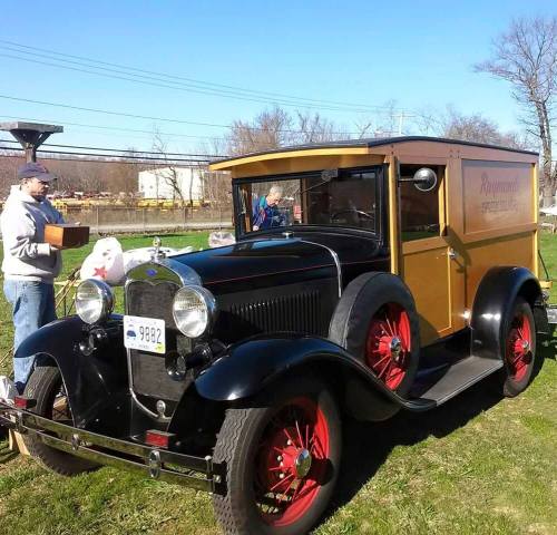A vintage truck with a wooden cargo area parked on grass, with two people nearby, one holding a box.