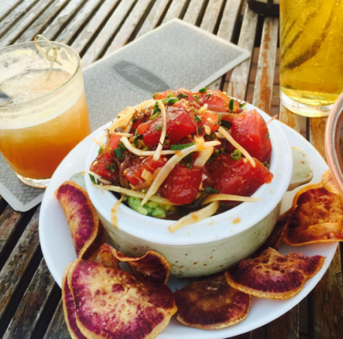 A bowl of vibrant tomato salad on guacamole, garnished with crispy toppings, served with colorful vegetable chips and a drink.