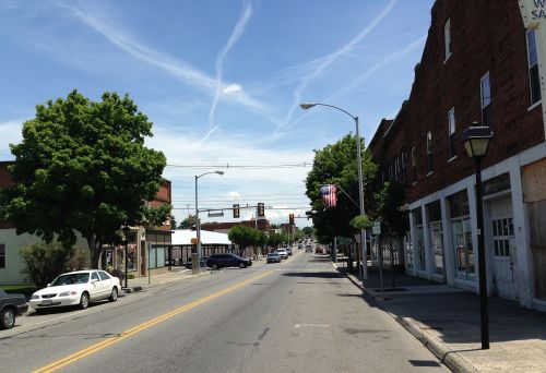 A quiet street scene with trees, buildings, and traffic lights under a blue sky with wispy clouds.