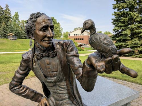 A bronze statue of a man holding a small bird on his hand, set in a park with trees and a building in the background.