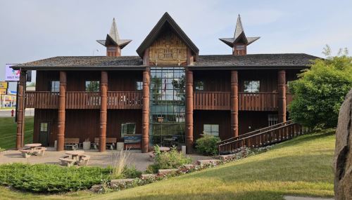 A two-story wooden building with a unique roof design, surrounded by greenery and flower beds.