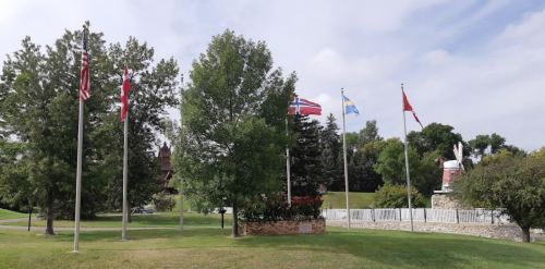 A park scene featuring several flags, including the U.S., Norway, and Canada, with trees and a windmill in the background.