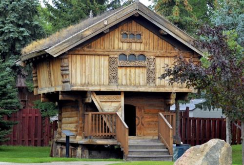 A rustic wooden house with intricate carvings, a thatched roof, and a staircase, surrounded by trees and a red fence.