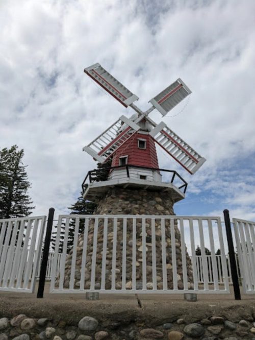 A red and white windmill atop a stone base, surrounded by a white fence under a cloudy sky.
