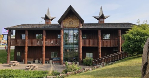 A unique wooden building with a peaked roof and large windows, surrounded by greenery and picnic tables.