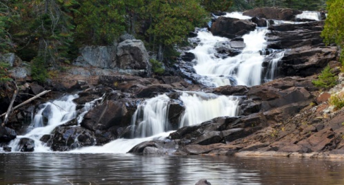 A serene waterfall cascades over rocky terrain, surrounded by lush greenery and reflecting in a calm pool below.