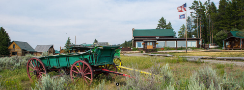 A green wagon sits in a grassy area with rustic buildings and flags in the background, surrounded by trees.