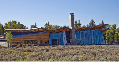 Modern building with large glass windows and wooden accents, surrounded by greenery and shrubs under a clear blue sky.