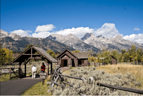 A rustic building near a path, surrounded by mountains and greenery under a clear blue sky.