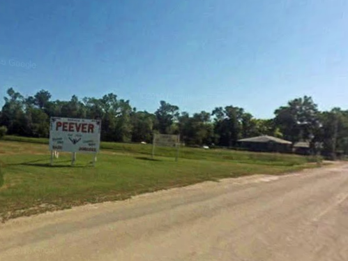 A rural road scene featuring a sign for "Peever" and a grassy area with trees in the background under a clear blue sky.