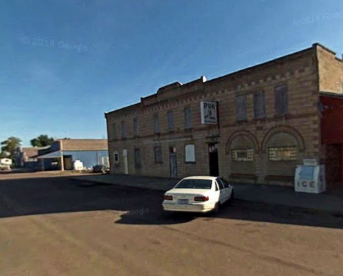 A street view of a brick building with arched windows, a parked white car, and a clear blue sky.