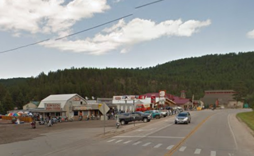 A small town street with shops, vehicles, and mountains in the background under a partly cloudy sky.