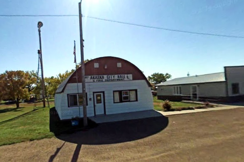 A small, white building with a red roof labeled "Akaska City Hall," surrounded by grass and trees.