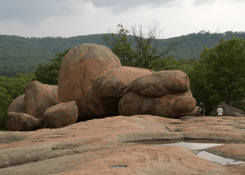 Large, weathered boulders on a rocky landscape, surrounded by greenery and a cloudy sky. People are visible in the background.