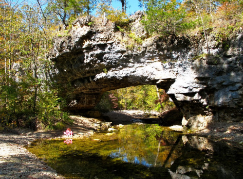 A natural stone arch over a clear stream, surrounded by trees and autumn foliage.
