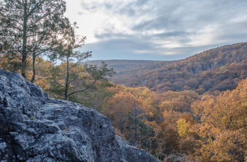 A scenic view of a colorful autumn landscape with rolling hills and trees, framed by rocky outcrops under a cloudy sky.