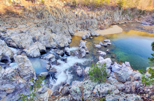 A serene river flows through rocky terrain, surrounded by trees and a calm pool of water.