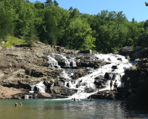 A scenic waterfall cascades over rocks, with people swimming in the clear water below, surrounded by lush greenery.