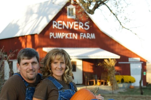 A couple in overalls sits with a pumpkin in front of a red barn labeled "Renyers Pumpkin Farm."
