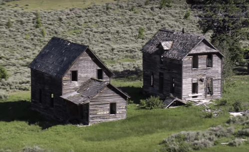 Two dilapidated wooden houses sit in a grassy field, surrounded by sparse vegetation and trees.