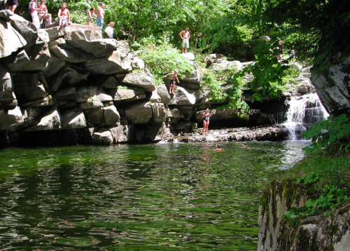 A natural swimming area with people on rocks and in the water, surrounded by lush greenery and a small waterfall.