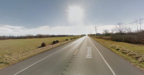 A straight road stretches into the distance, flanked by fields and hay bales under a bright sun.