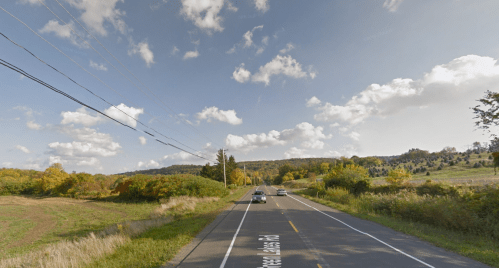 A scenic road lined with trees and fields, under a blue sky with fluffy clouds. Cars are visible on the road.