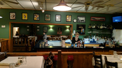 A cozy restaurant bar with a green wall, shelves of drinks, and a bartender serving customers at the counter.