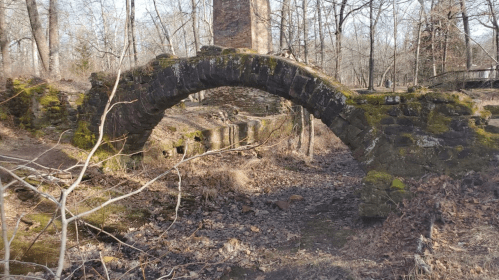 A moss-covered stone arch bridge over a dry creek bed, surrounded by bare trees and rocky terrain.