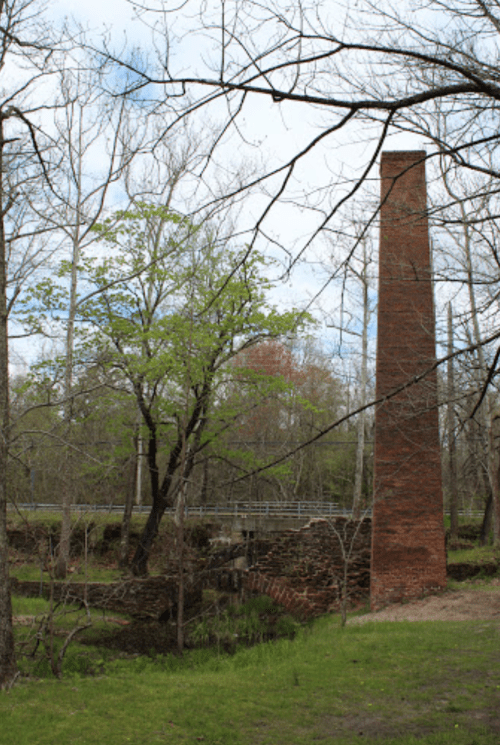 A tall brick chimney stands among trees, with a stone structure and a bridge in the background under a cloudy sky.