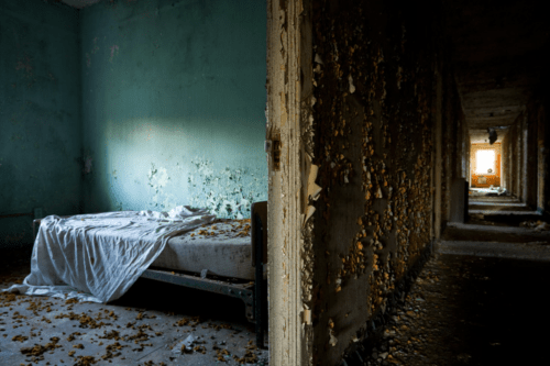 Abandoned room with a disheveled bed, peeling walls, and debris scattered on the floor, illuminated by soft light.