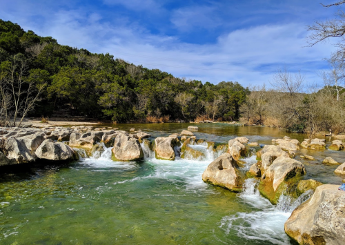 A serene river scene with flowing water over rocks, surrounded by lush greenery and a clear blue sky.
