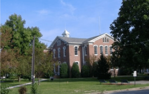 Historic brick building with large windows, surrounded by trees and green space under a clear blue sky.