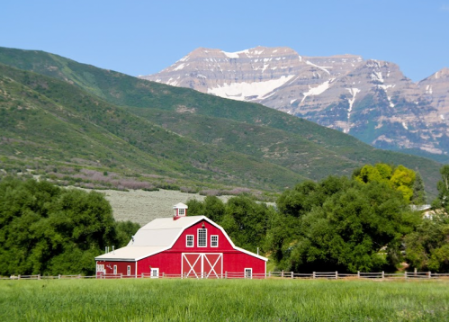 A red barn stands in a green field, with mountains and blue sky in the background.