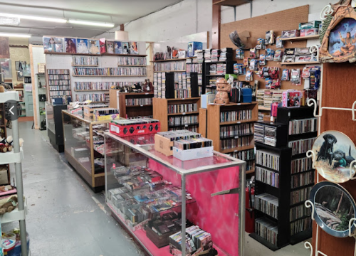 A cluttered store interior filled with shelves of CDs, DVDs, and collectibles, with a display case in the foreground.