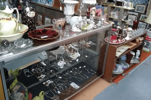 Display case filled with jewelry and decorative items, surrounded by various collectibles and home decor in a shop.