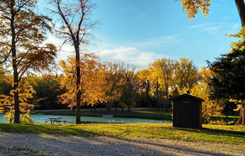 A serene park scene with autumn trees, a small shed, and a pond under a clear blue sky.