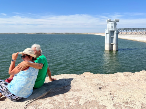 A couple sits together on a rocky shore by a lake, enjoying the view and sunshine on a clear day.