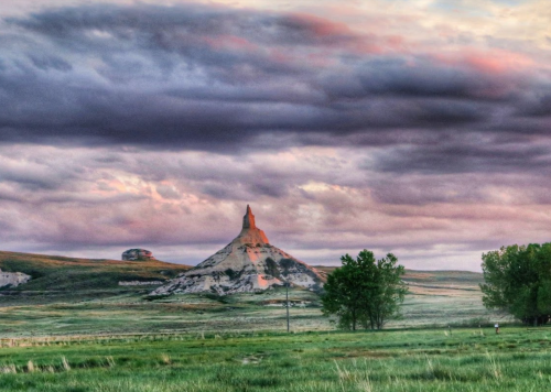 A dramatic landscape featuring a towering rock formation under a colorful sky, surrounded by green fields and trees.