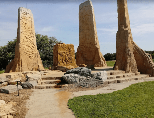 Four tall stone pillars with carvings, surrounded by grass and rocks, under a clear blue sky.