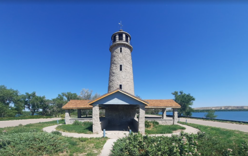 A tall stone lighthouse with a wooden roof, surrounded by greenery and a clear blue sky, near a body of water.