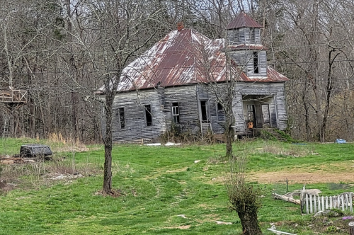 Abandoned, weathered house with a rusty roof, surrounded by overgrown grass and trees.