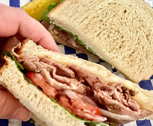 A hand holding a sandwich with layers of meat, lettuce, and tomato, with another sandwich in the background.