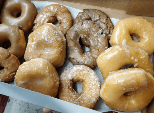 A box of assorted donuts, including glazed, powdered, and cake varieties, arranged neatly on a white paper liner.
