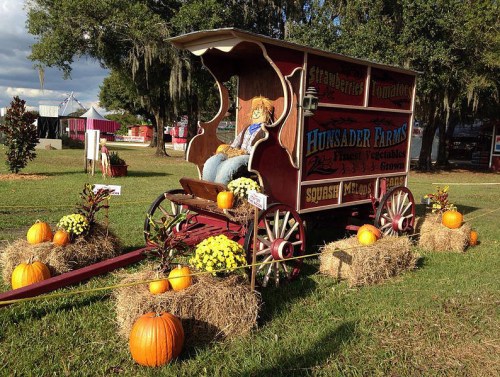 A colorful farm cart decorated with pumpkins, hay bales, and flowers, set in a grassy area under a blue sky.