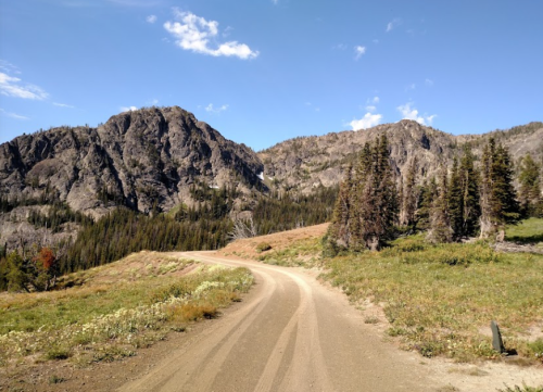 A winding dirt road leads through a mountainous landscape with trees and blue skies.